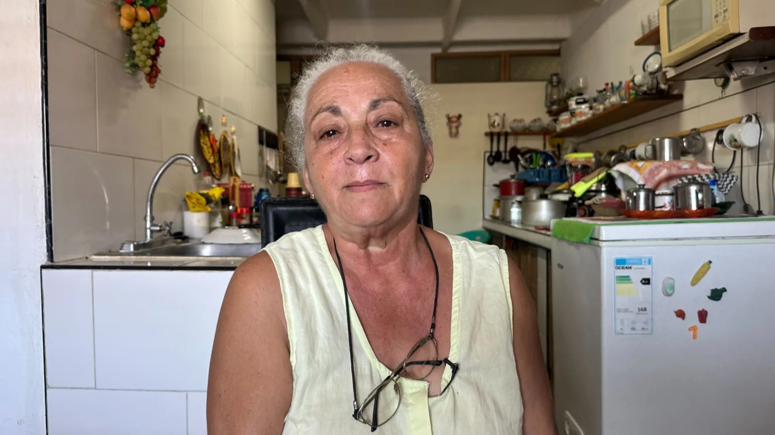 BBC A woman wearing a yellow, short-sleeve top and with glasses on a string around her neck sits in front of a kitchen 