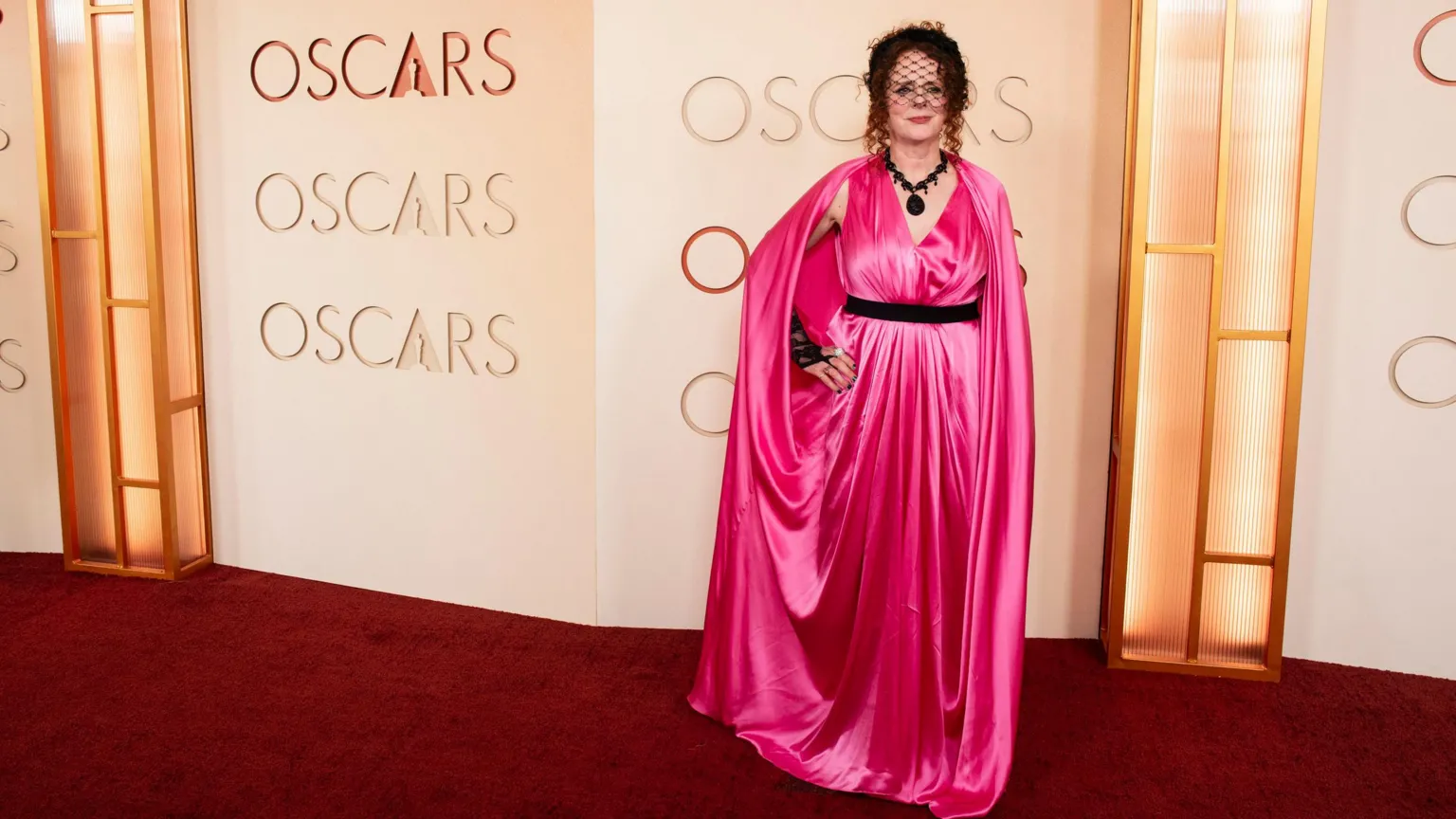 Getty Images Maggie is bright pink long satin dress, black belt, black necklace & little veil hat. Posing behind the word Oscars.