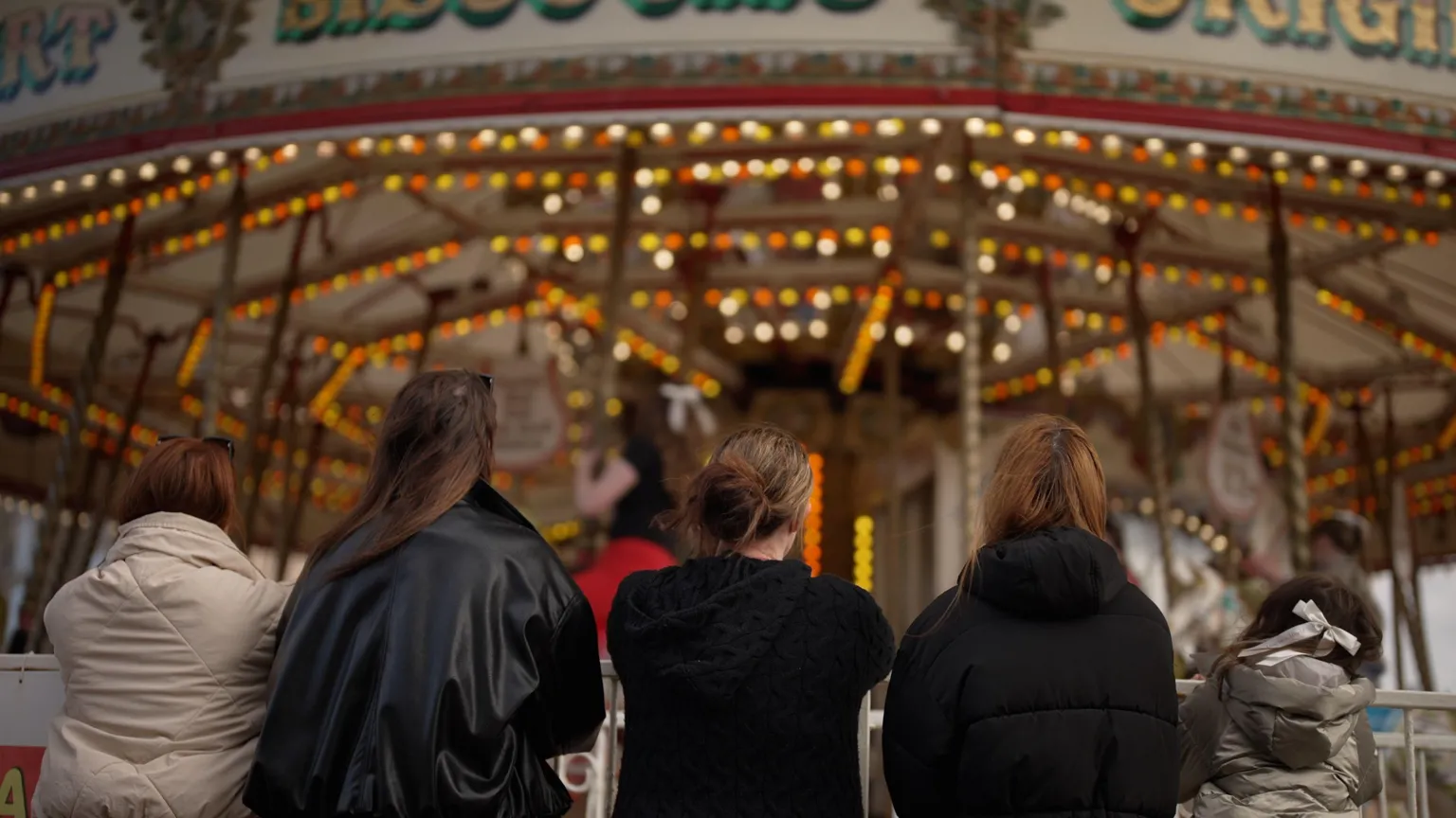 Brijesh Patel/BBC Four women stand in a row, with their backs to the camera, looking at a funfair carousel going round